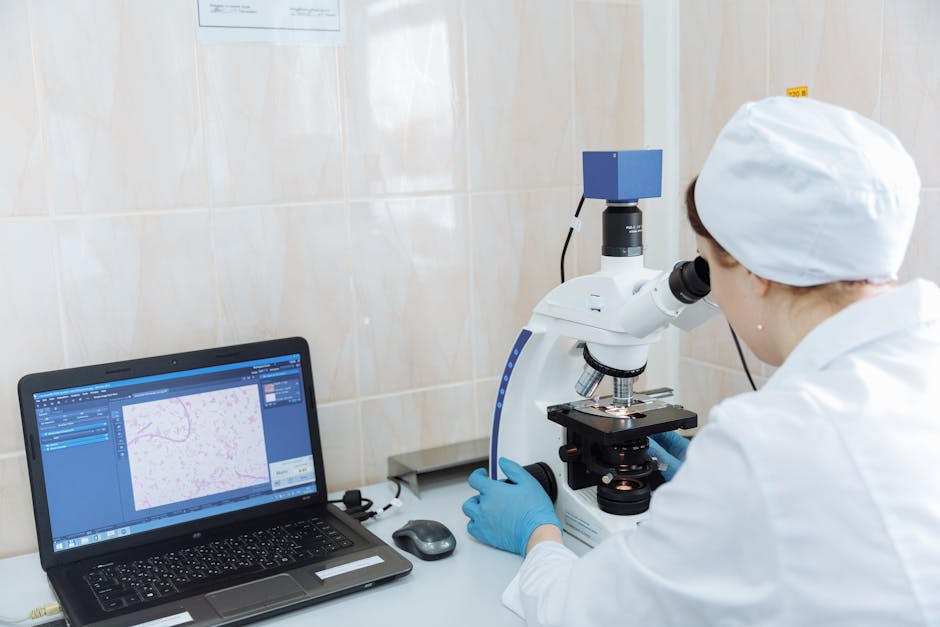 Scientist in lab coat using microscope and laptop in a laboratory setting.